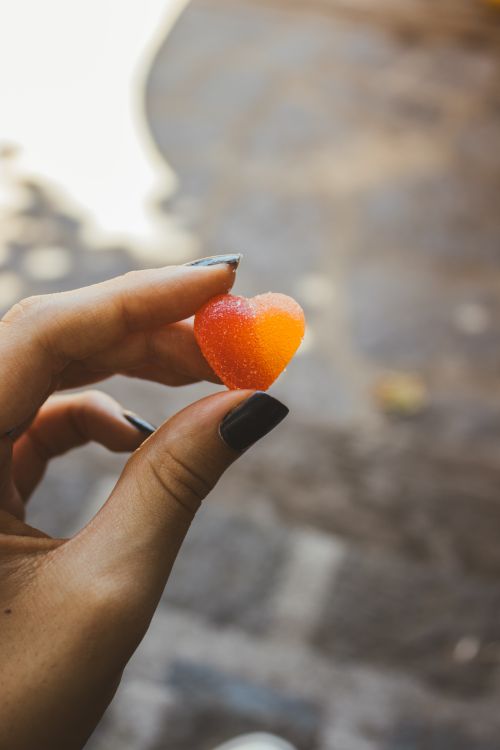 Free A close-up of a hand holding a heart-shaped gummy candy with a blurred outdoor background. Stock Photo