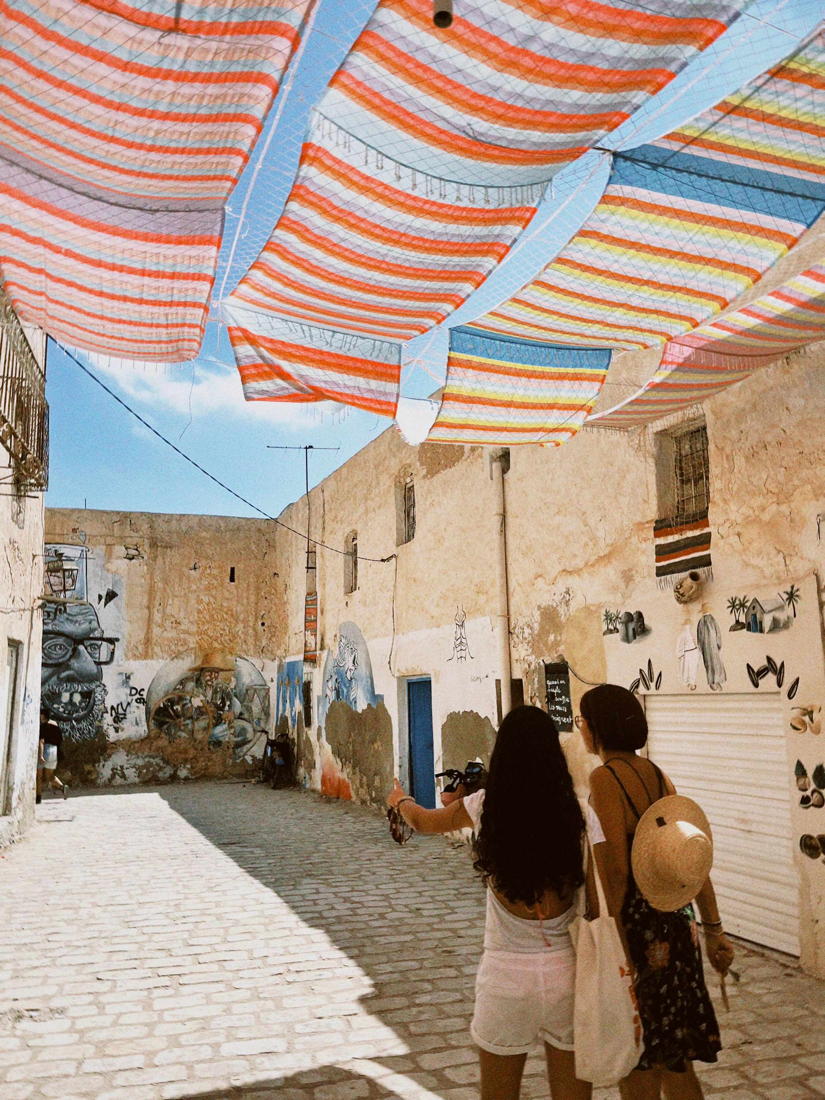 Two women walk under colorful canopies in the artistic streets of Djerba, Tunisia, capturing summer's essence.