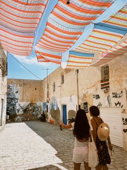Two women walk under colorful canopies in the artistic streets of Djerba, Tunisia, capturing summer's essence.
