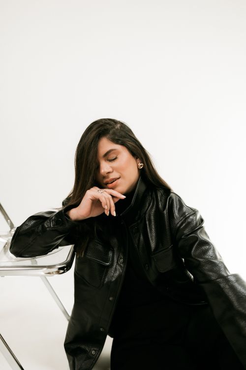 Free Fashionable woman in black leather jacket seated against minimalist background. Stock Photo