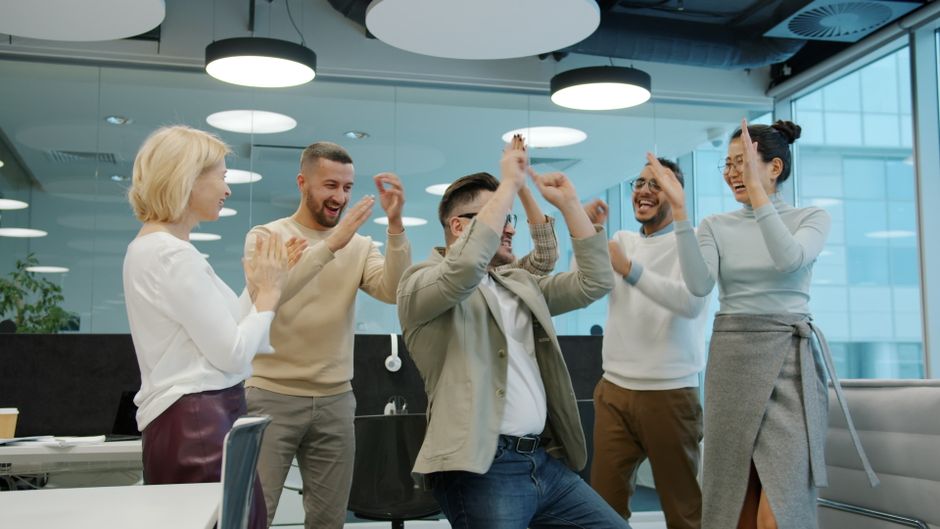 Un grupo diverso de compañeros celebrando juntos en un entorno de oficina moderno, chocando las manos y sonriendo.