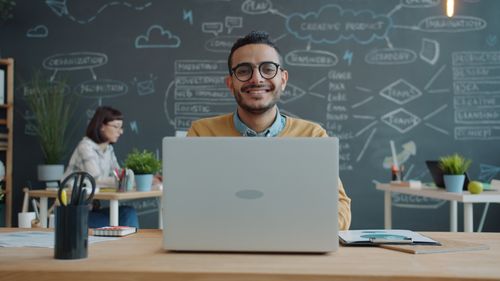 Free Smiling professional working on laptop in modern, creative workspace with chalkboard wall. Stock Photo