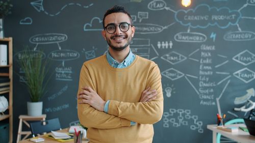 Free Smiling young man in casual attire stands confidently in a modern office with a chalkboard background. Stock Photo