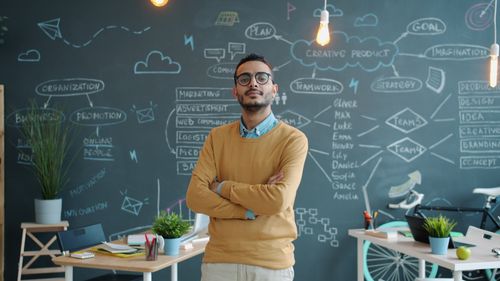 Free Entrepreneur standing confidently in a modern office with a creative chalkboard background. Stock Photo