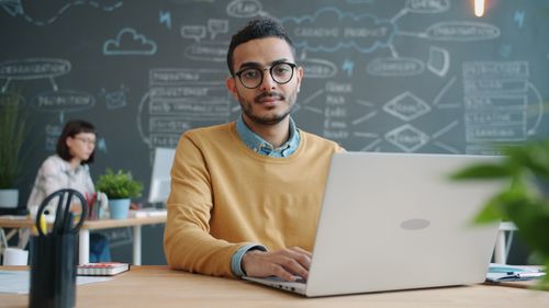 Free Young adult working on laptop in a modern office with creative chalkboard background. Stock Photo