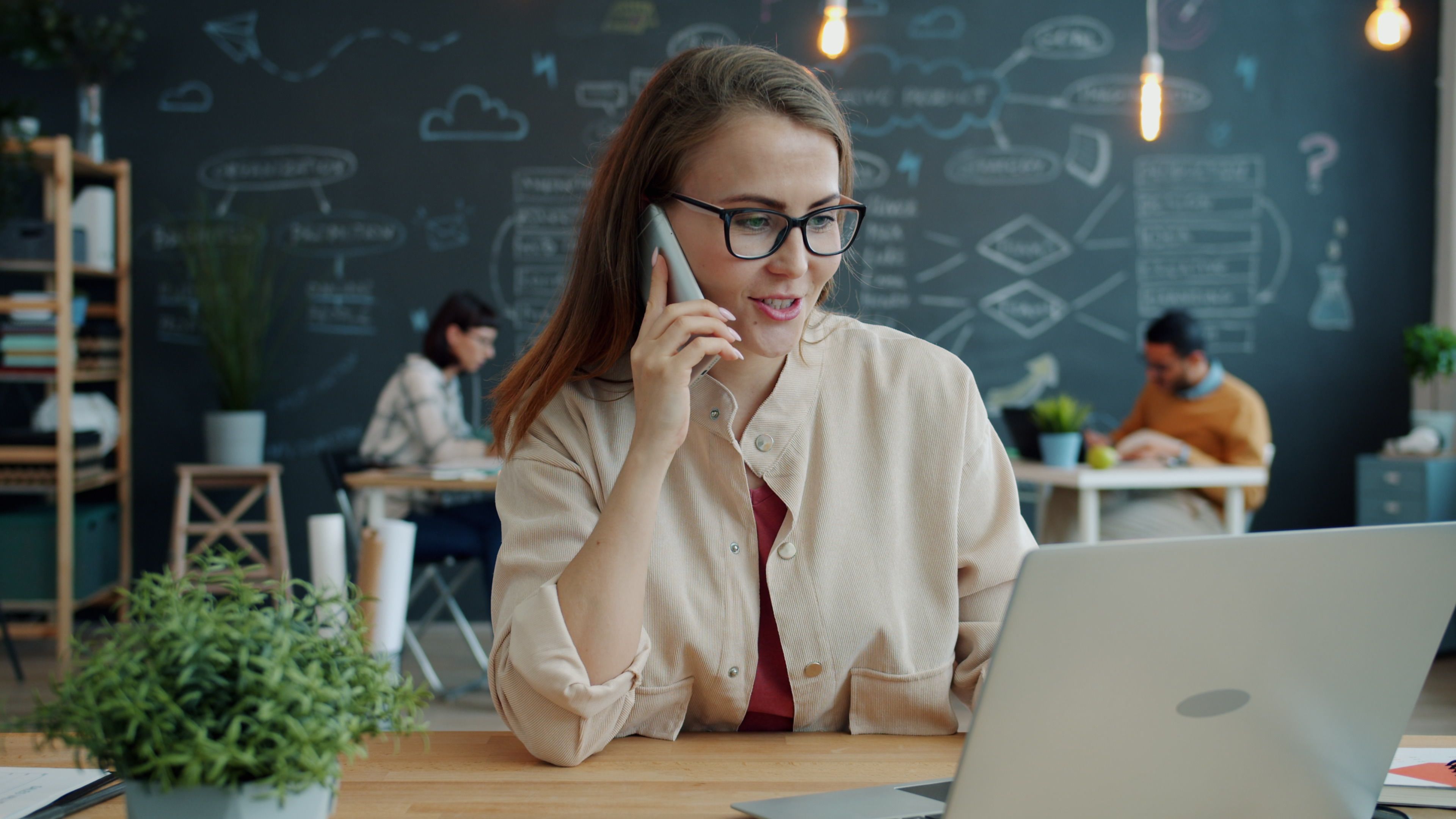 Young woman on phone and laptop in modern office with creative blackboard.