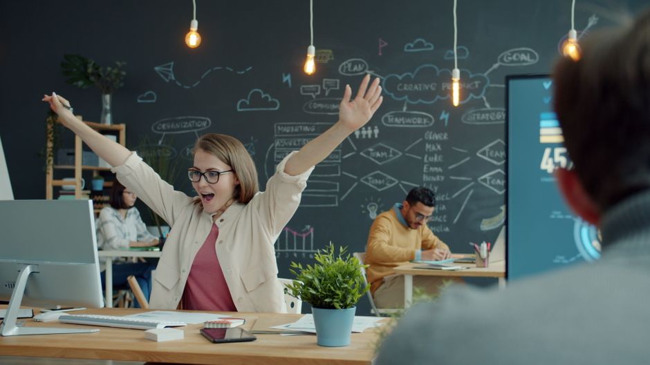 Young woman excitedly celebrates success in a modern office setting, arms raised with joy.