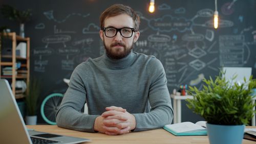Free Young professional sitting at desk in modern office with chalkboard wall, looking focused. Stock Photo
