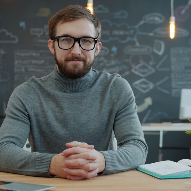 Mann mit Bart und Brille sitzt am Schreibtisch vor einer Tafel