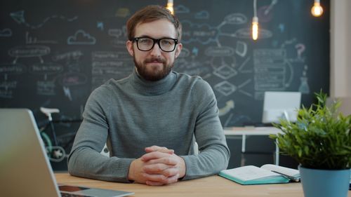 Free Confident professional sitting at a desk in a modern office with a creative chalkboard background. Stock Photo