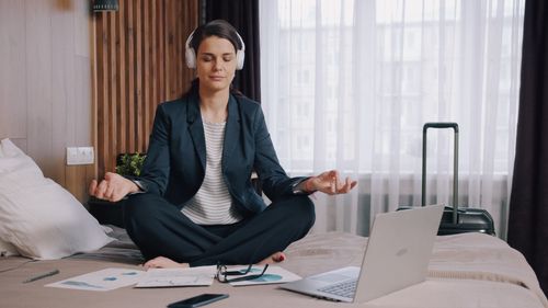 Free Businesswoman seated on a bed meditating with headphones and laptop in hotel room. Stock Photo