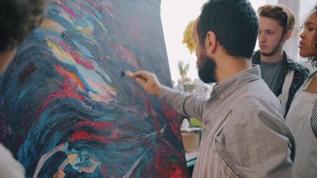 A diverse group of young adults watches an artist painting a vibrant abstract artwork in a studio.
