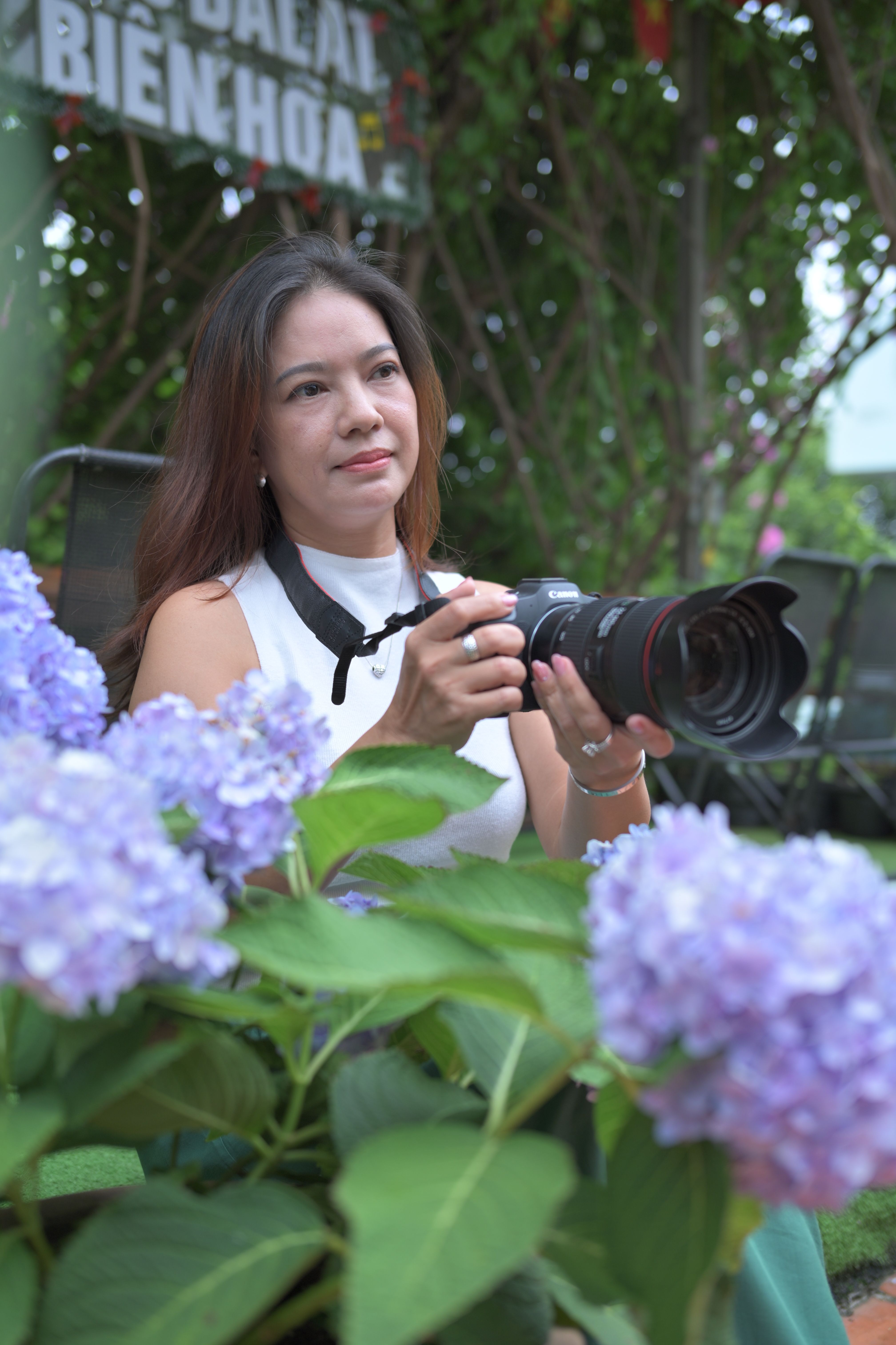 A woman photographer holding a camera surrounded by vibrant purple flowers in a garden setting.
