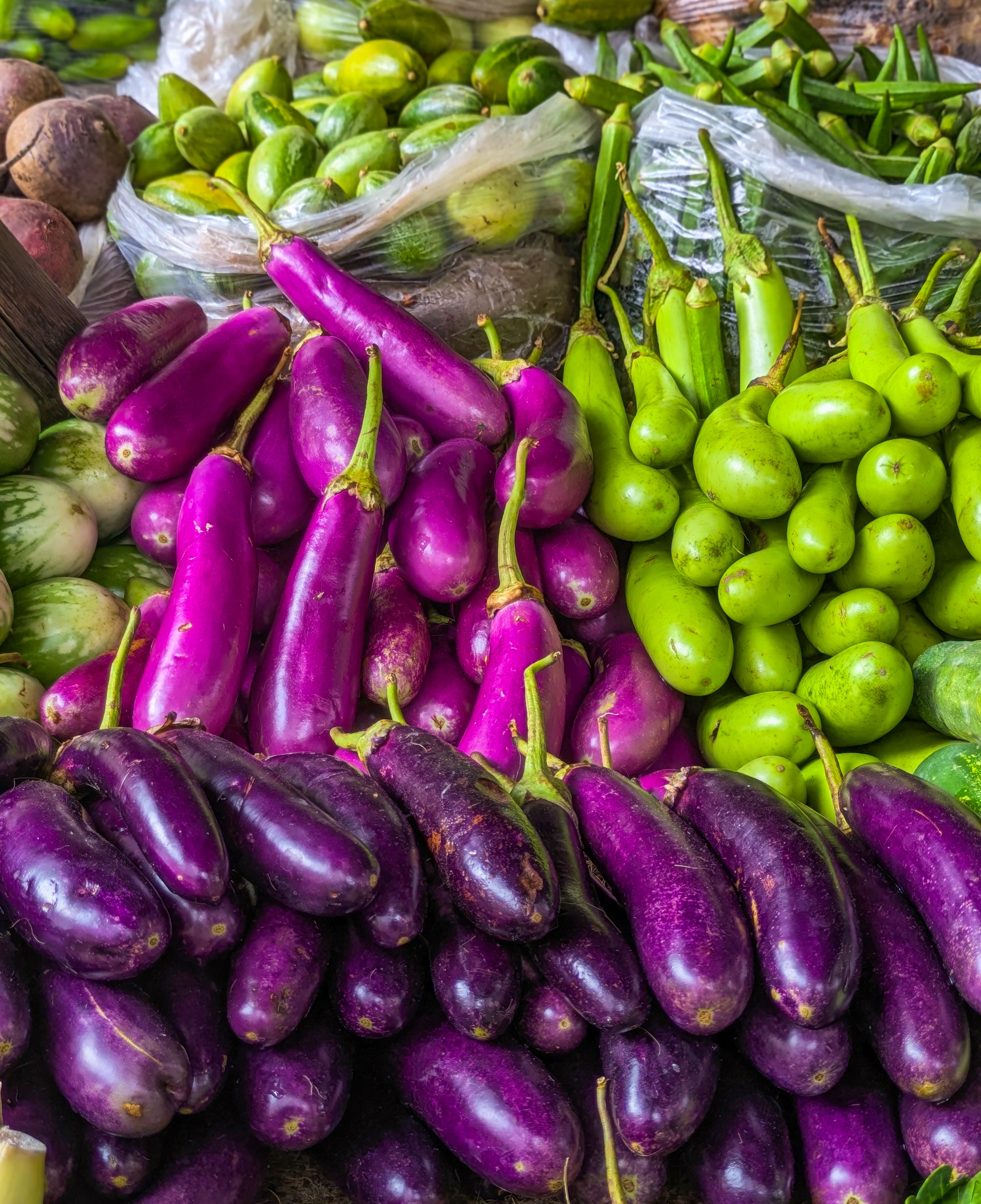 Heaps of purple brinjals beside green gourds and okra on a vegetable market stall.
