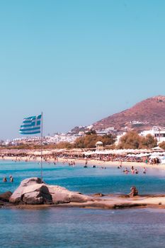 Vibrant summer beach in Greece with people relaxing, Greek flag waving, and scenic hills.