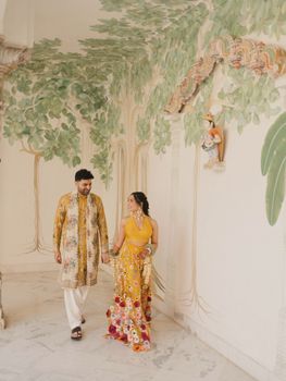 A stylish couple walks hand in hand in vibrant traditional attire against an artistic leafy backdrop.