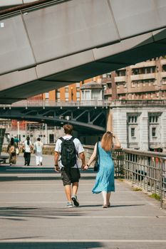 A couple walking hand-in-hand near Zubizuri Bridge in Bilbao, capturing a sunny summer day.