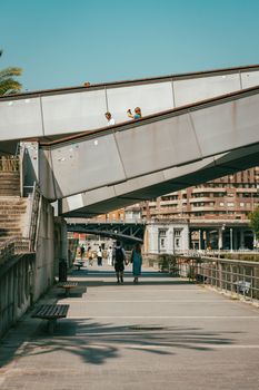 People walking under a modern bridge in Bilbao, Spain, on a sunny day.