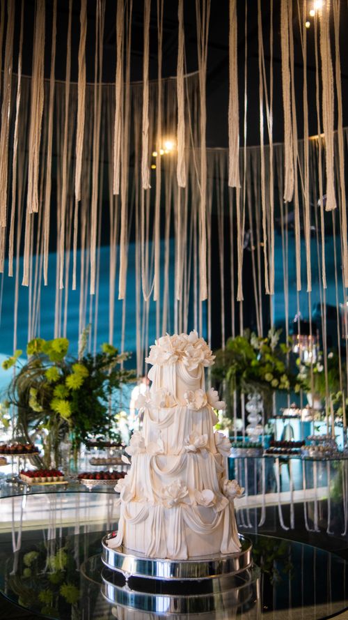 Free Exquisite multi-tiered wedding cake with intricate floral drapes on display under decorative strands. Stock Photo