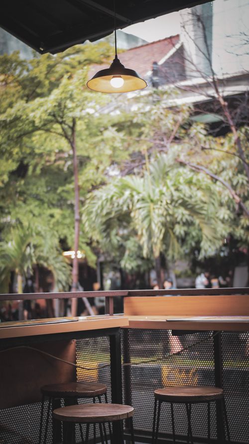 Free Charming outdoor cafe setting with empty stools under a vintage street lamp, surrounded by lush greenery. Stock Photo