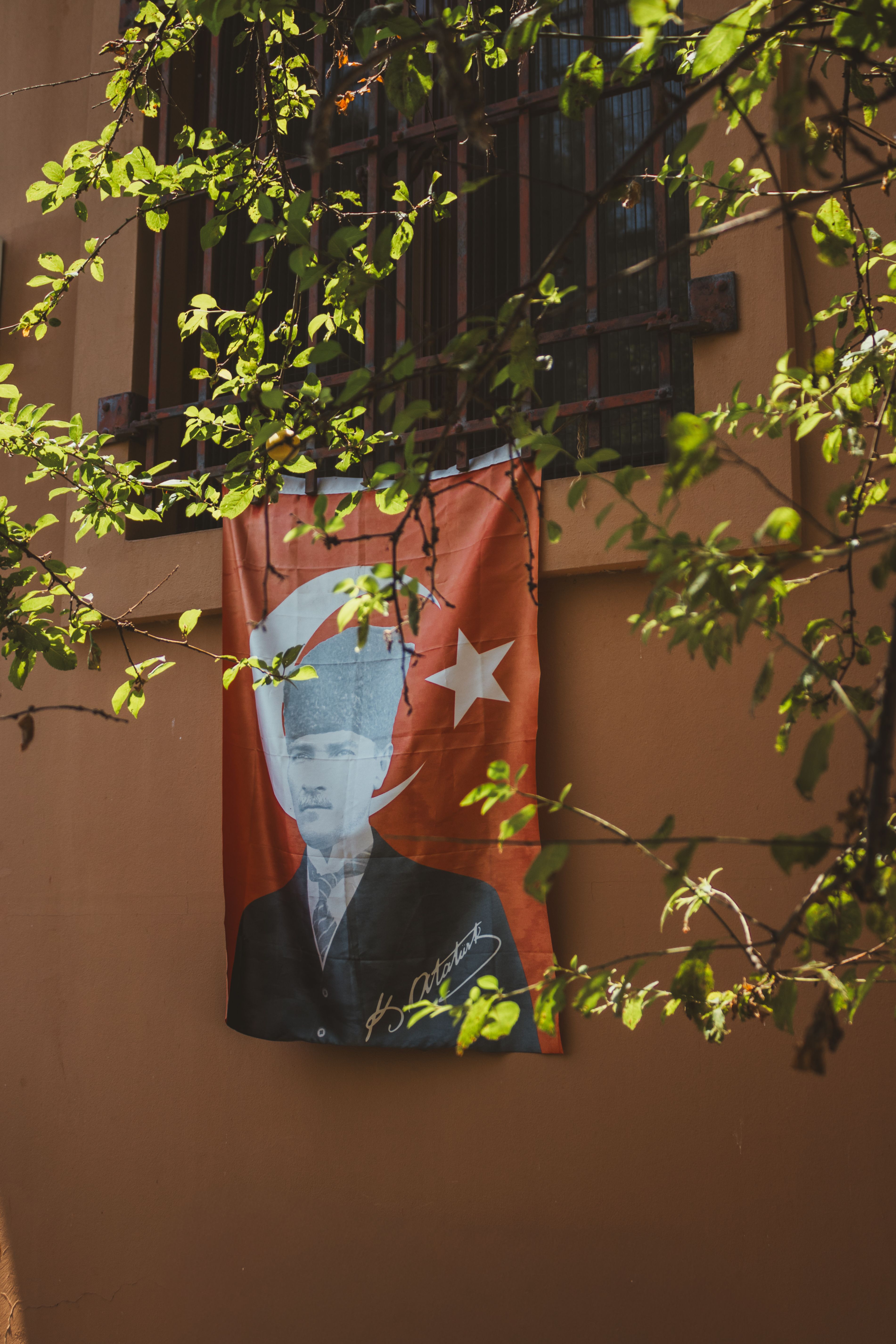 A Turkish flag with a portrait hangs outside a building window, framed by tree branches.
