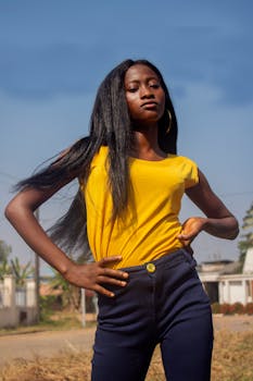Confident young African American female with long black hair in casual outfit standing in countryside with hands on waist and looking at camera