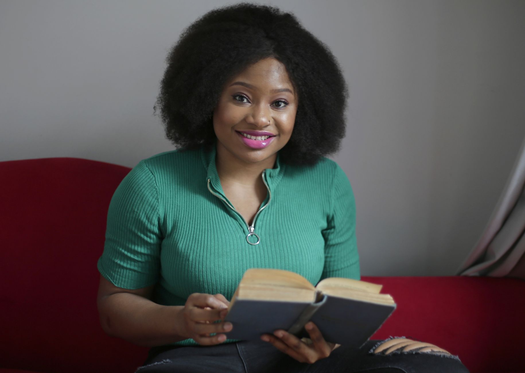 African American woman with afro hair sitting on a couch and reading a book with a smile.