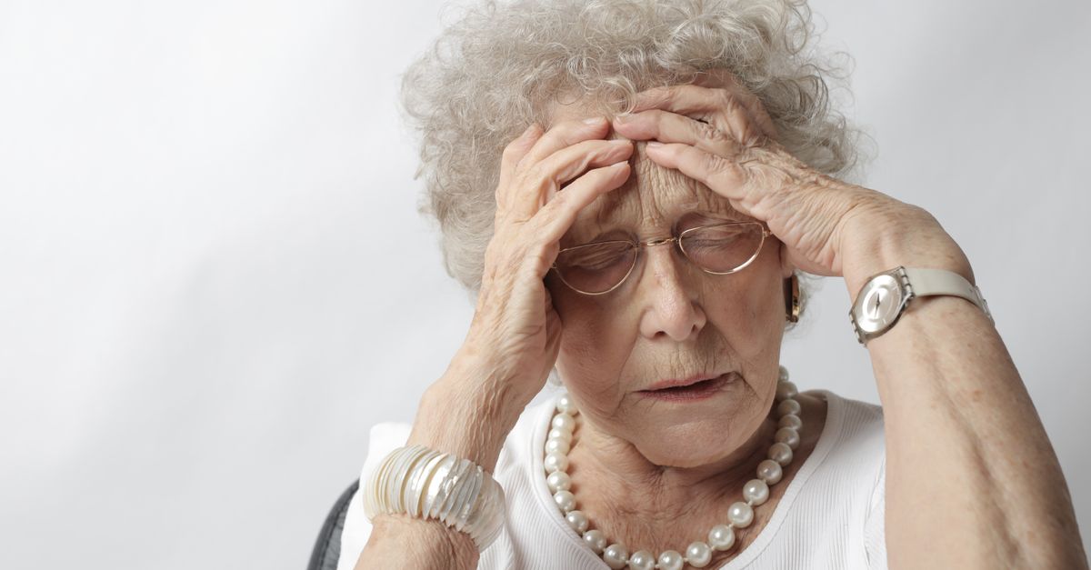 An elderly woman with her hands on her head, appearing stressed or in pain, illustrating Lewy Body Dementia Symptoms: A Complete Guide for Families โ 1-800-MEDIGAP, America's Trusted Toll-Free Number.