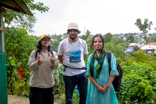 Three adults stand together smiling at a Bangladeshi refugee camp amidst lush greenery.
