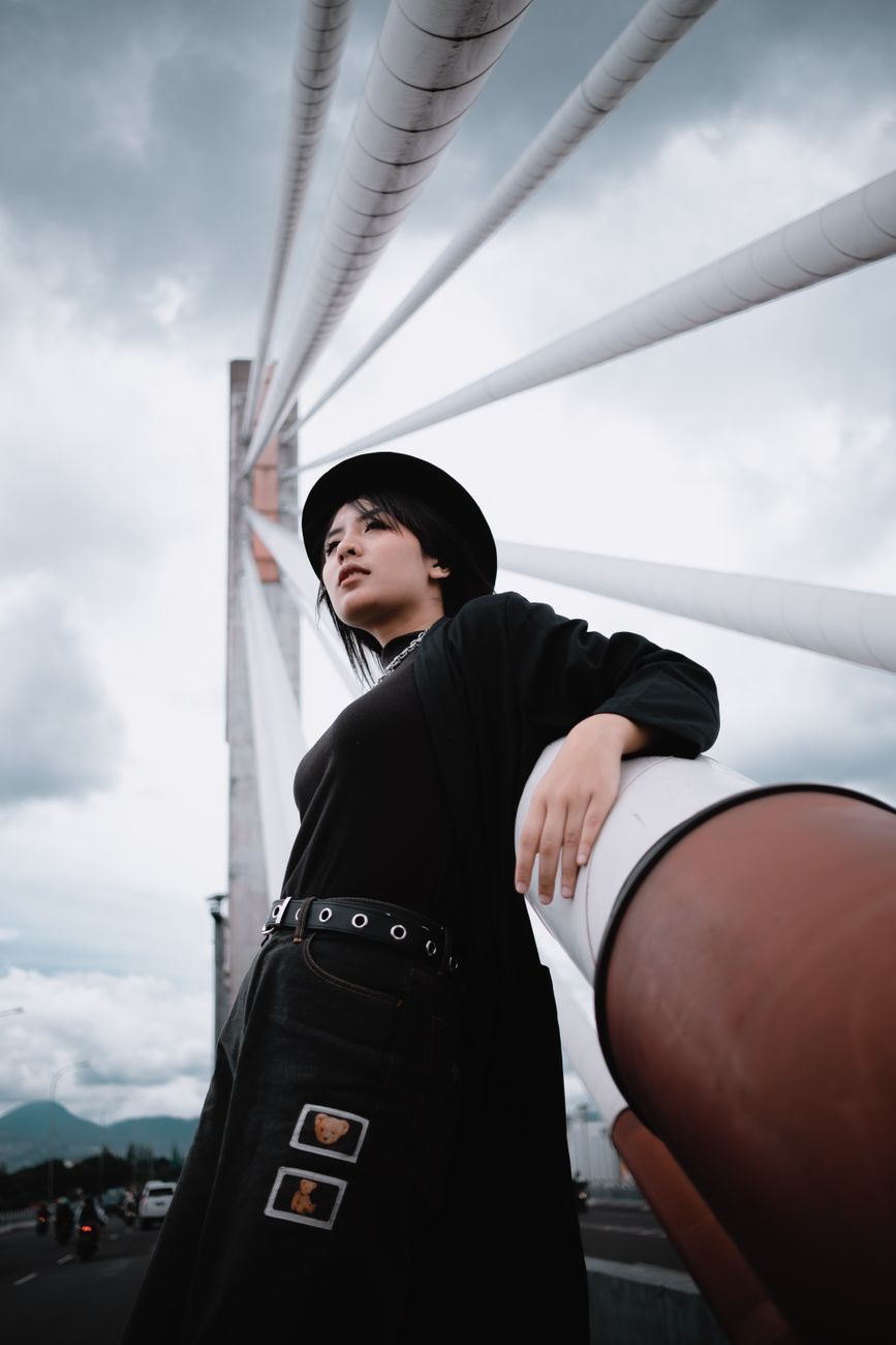 Elegant woman in black outfit posing against a modern bridge with a dramatic cloudy sky.