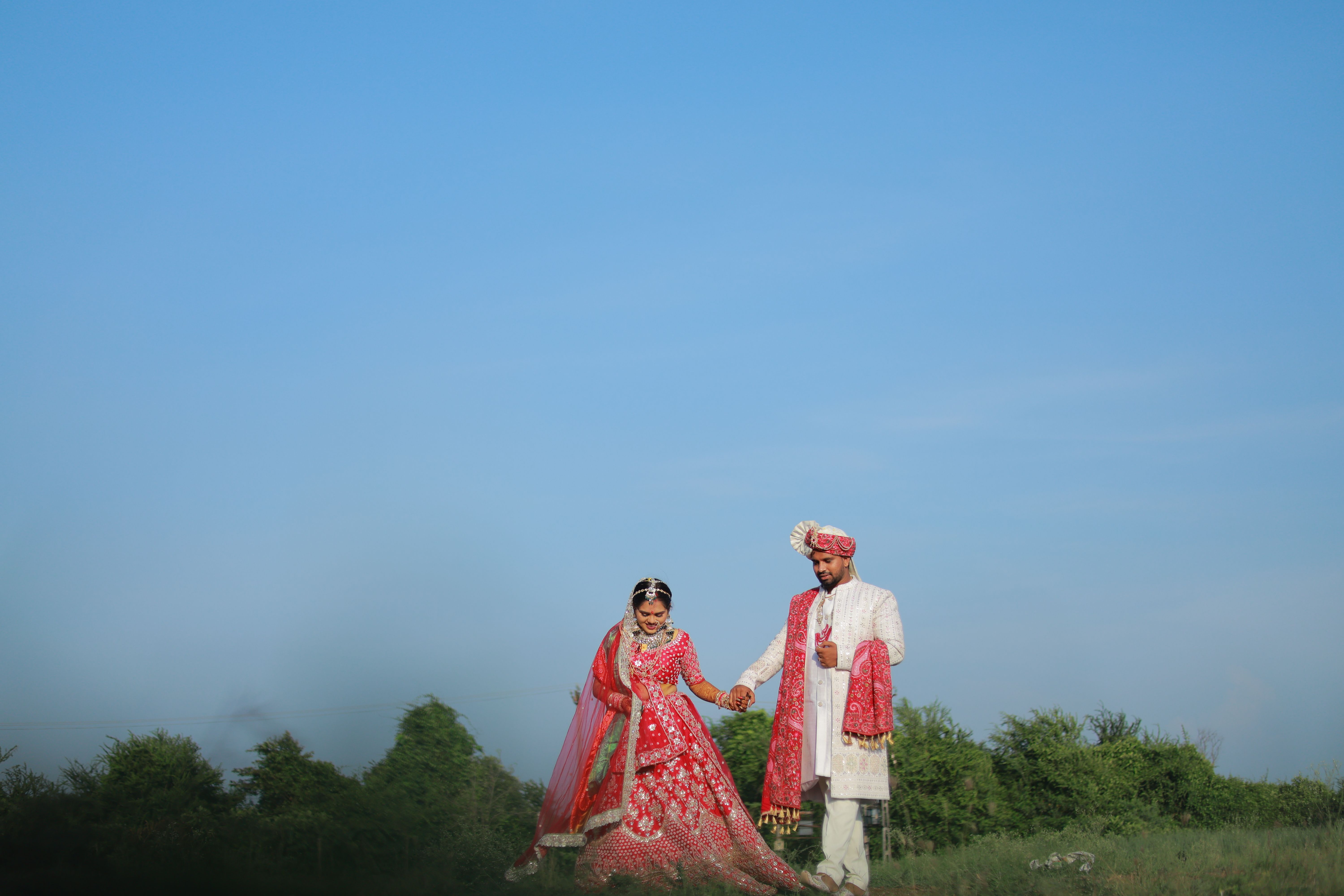 A joyful Indian bride and groom holding hands in traditional attire against a clear blue sky.