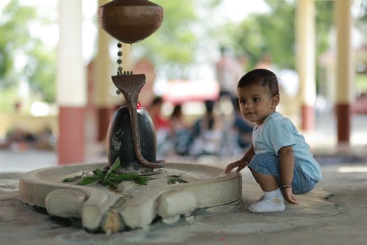 A cute baby, dressed casually, curiously exploring near a Shiva Lingam in a serene outdoor temple setting.