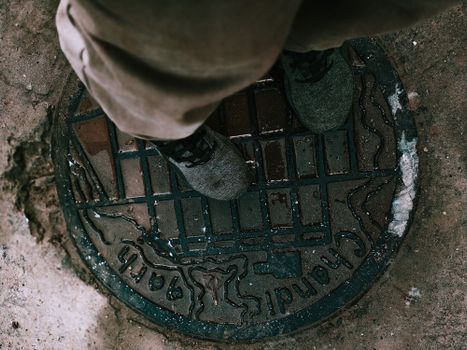 从上方看去，一个身穿靴子和裤子的匿名人正站在城市街道的破旧金属井盖上