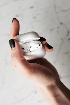 A hand with black nail polish holds a pair of wireless earbuds in a white case.