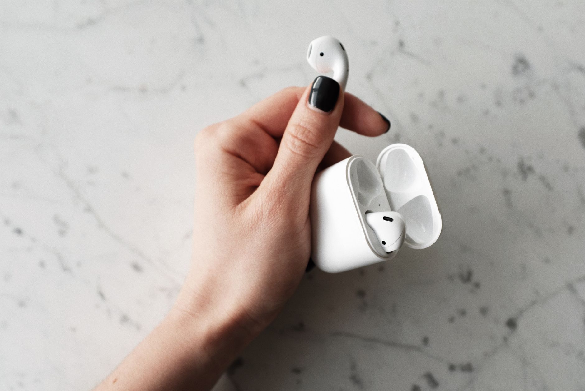 A close-up of a hand holding wireless earphones with black nail polish on a marble surface.