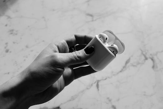 Black and white image of a hand holding wireless earbuds in a charging case on a marble surface.