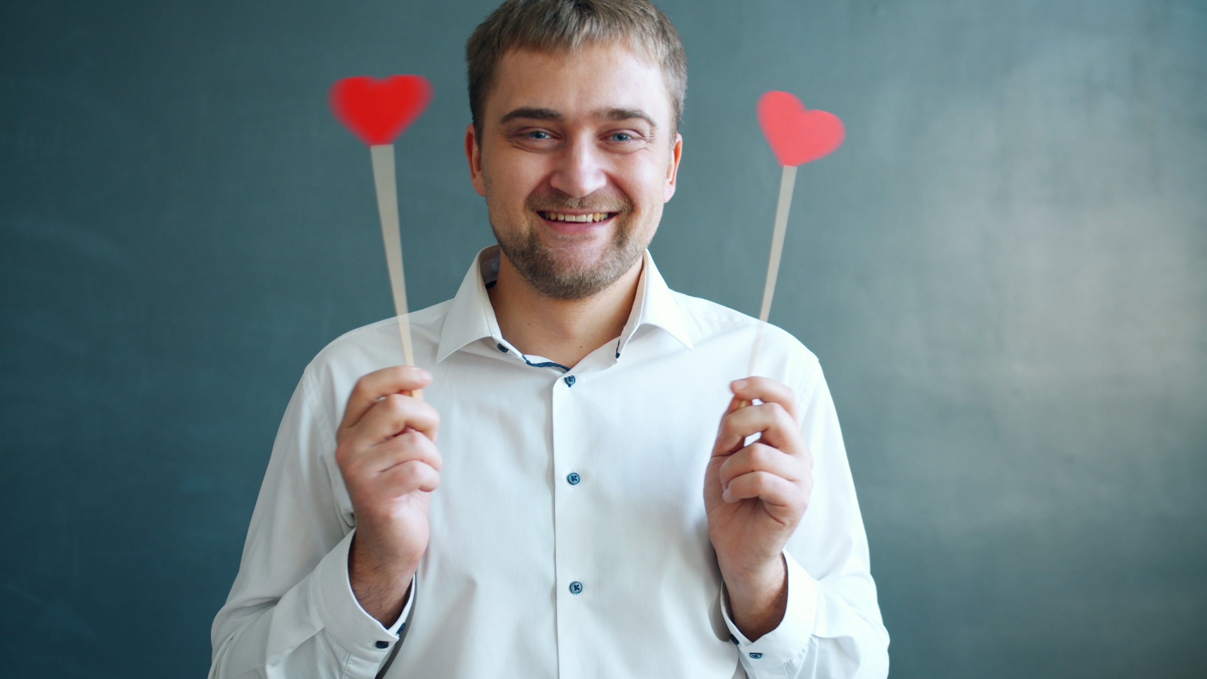 Cheerful man in shirt holding two heart-shaped stick decorations indoors.