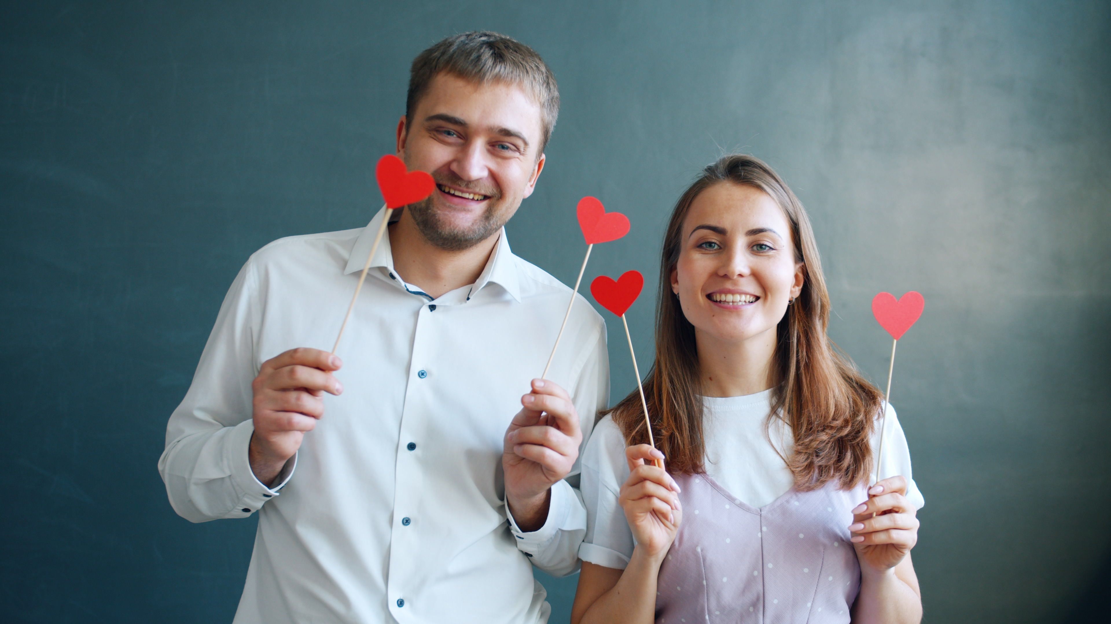 A cheerful couple poses with heart-shaped props against a plain backdrop.
