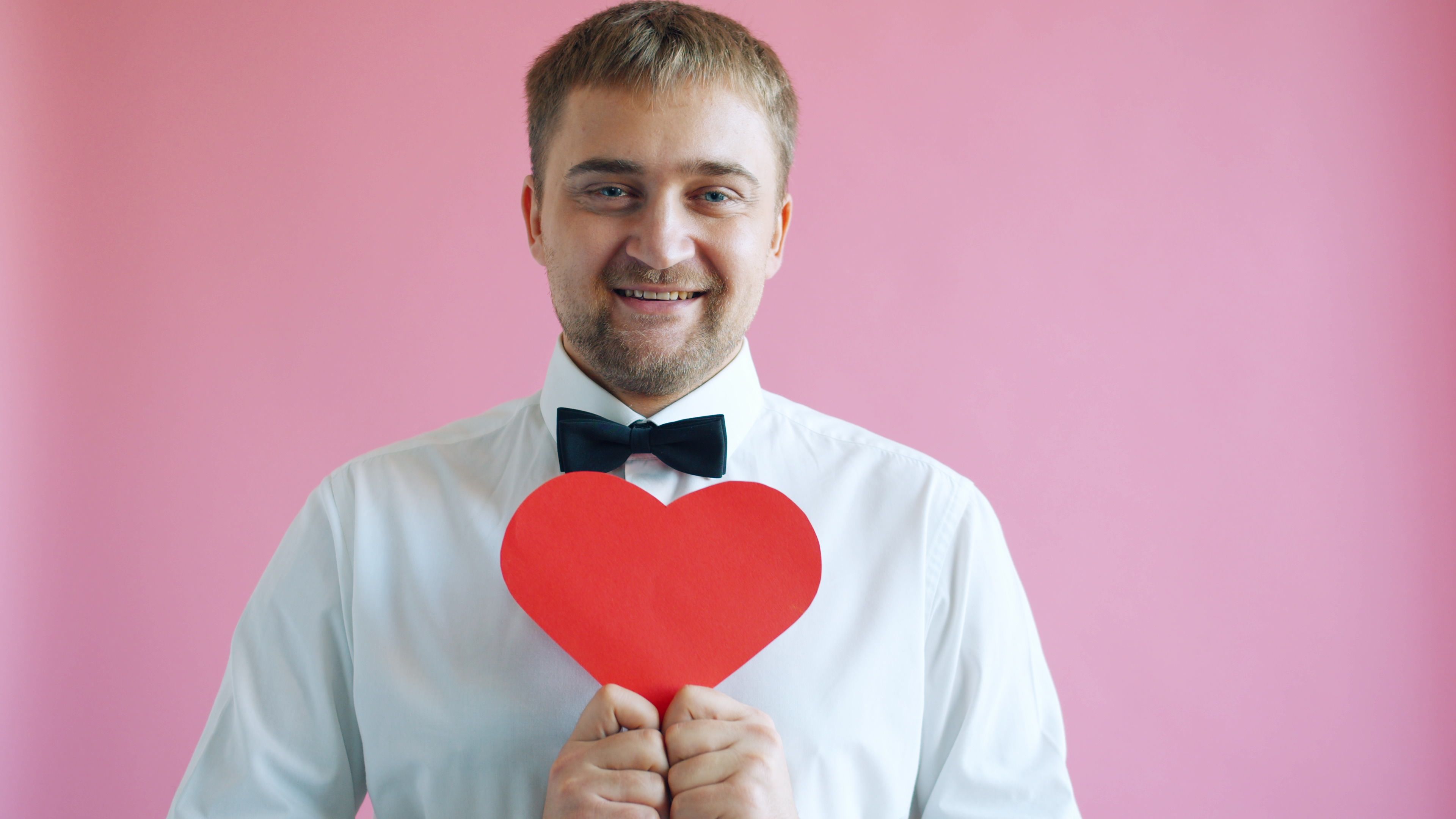 Portrait of a cheerful man in a white shirt holding a red heart shape against a pink backdrop.
