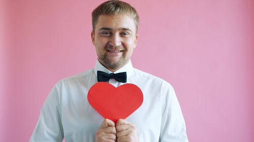Free Portrait of a cheerful man in a white shirt holding a red heart shape against a pink backdrop. Stock Photo