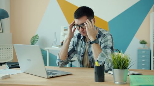 Free Young man in glasses looking stressed at his desk with a laptop and modern decor. Stock Photo