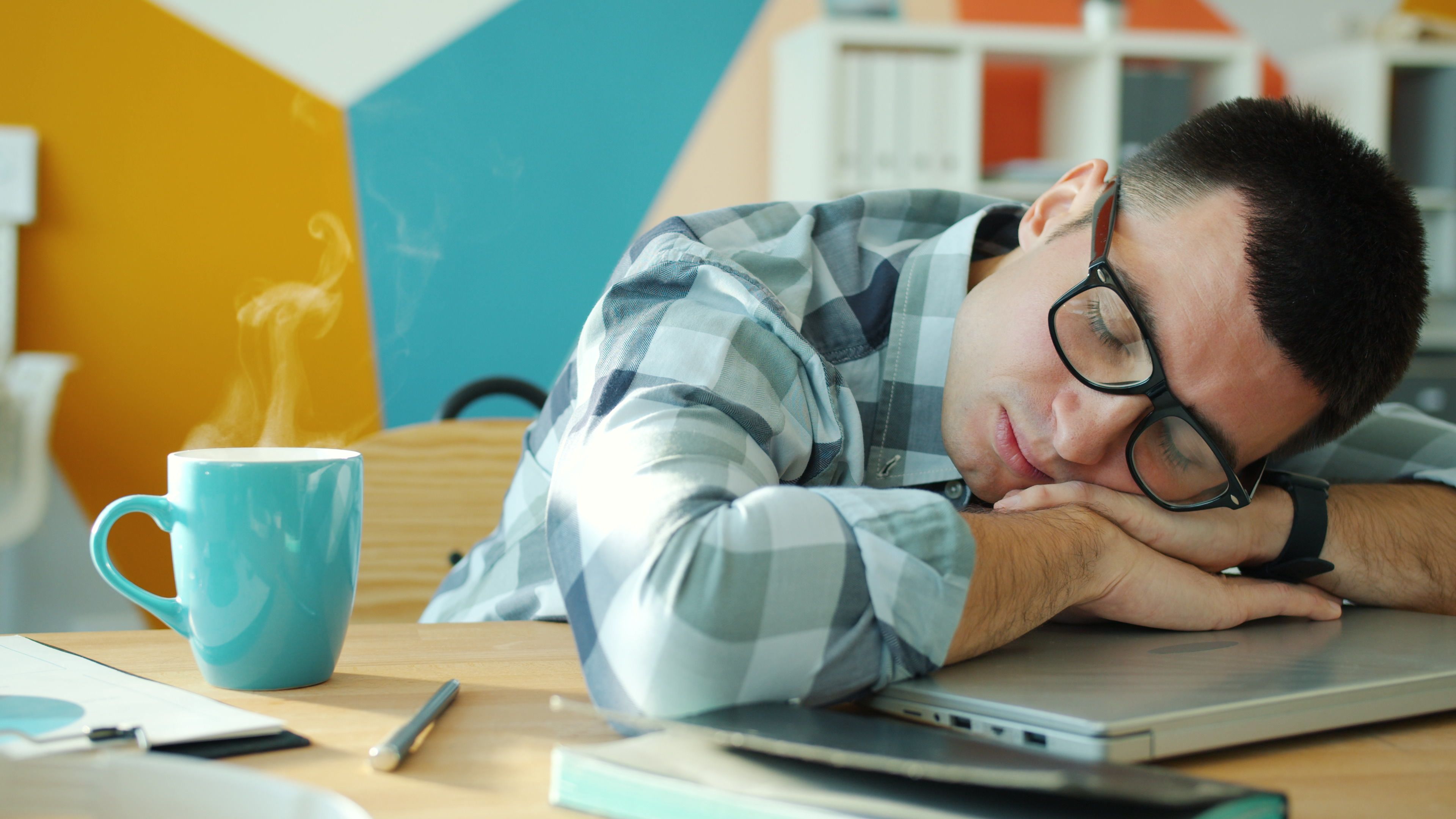 Tired man sleeping on laptop with coffee, showcasing workplace fatigue.