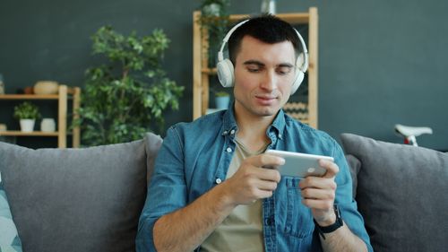 Free Casual young man relaxing on couch with headphones, playing games on smartphone. Stock Photo