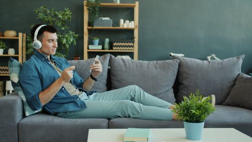 Free Young man relaxing on a couch with headphones, browsing on phone. Modern and cozy interior. Stock Photo