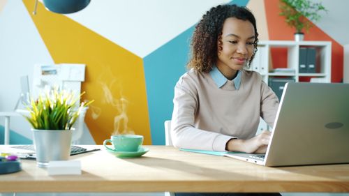 Free Young woman working on a laptop in a modern and colorful office setting. Stock Photo