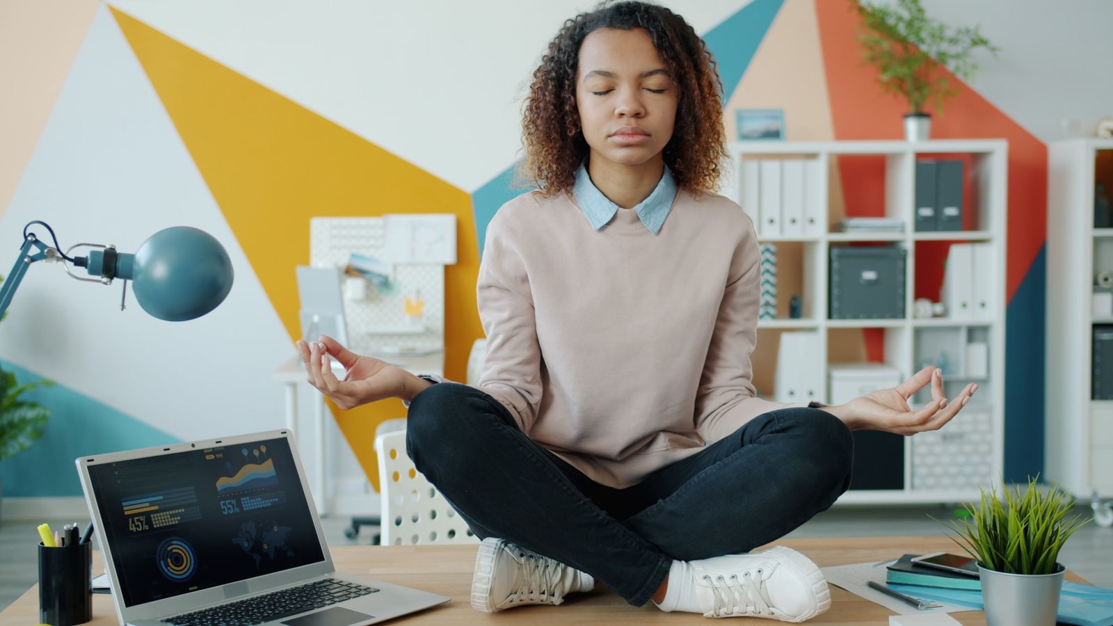 Relaxed young woman meditating cross-legged on desk in modern office.