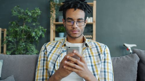 Free A young man wearing glasses uses a smartphone while seated on a sofa indoors. Stock Photo