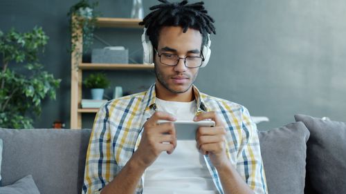 Free Young man with headphones playing on smartphone indoors, relaxing on sofa. Stock Photo