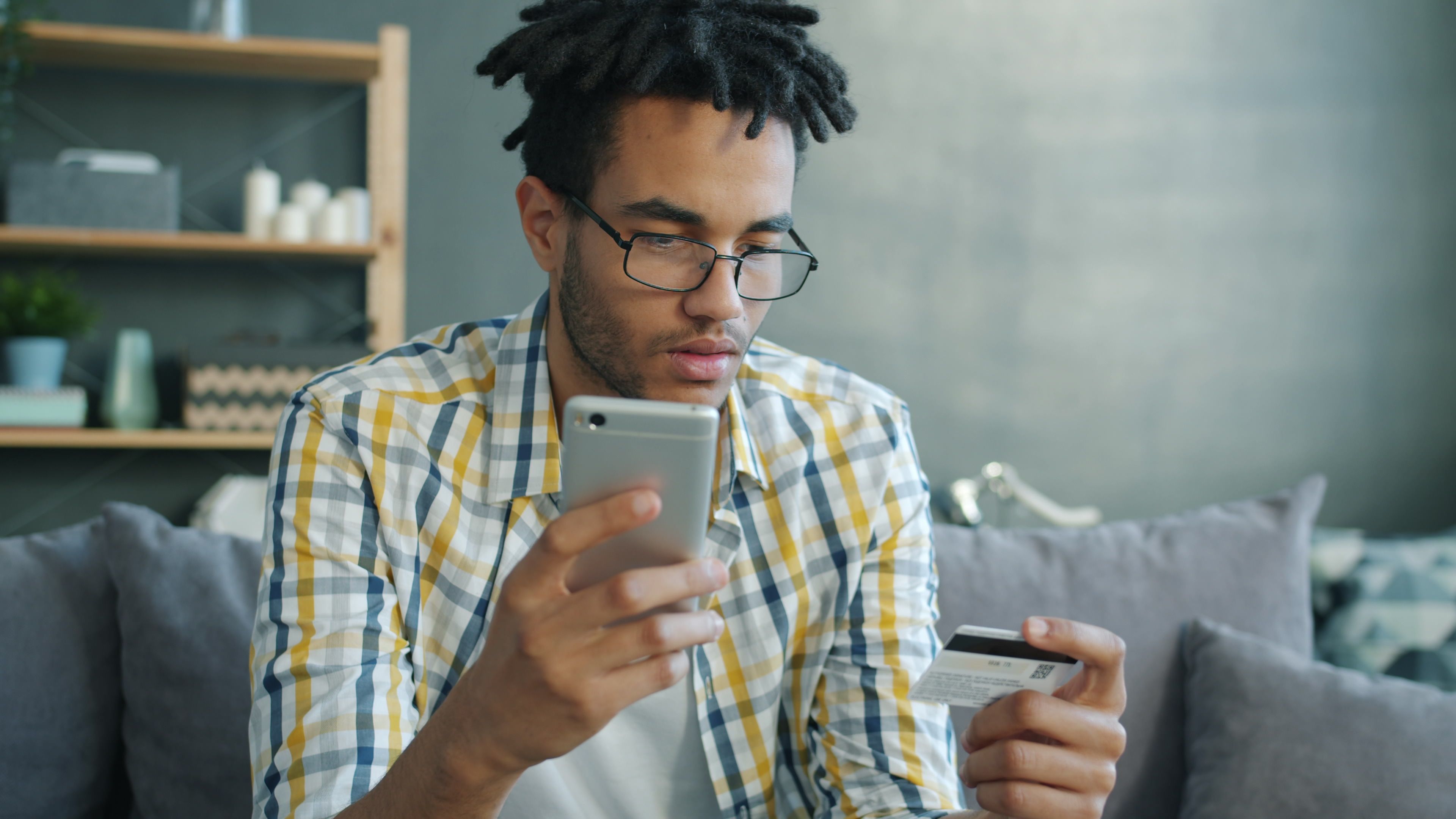 Young man shopping online with a smartphone and credit card in a living room.