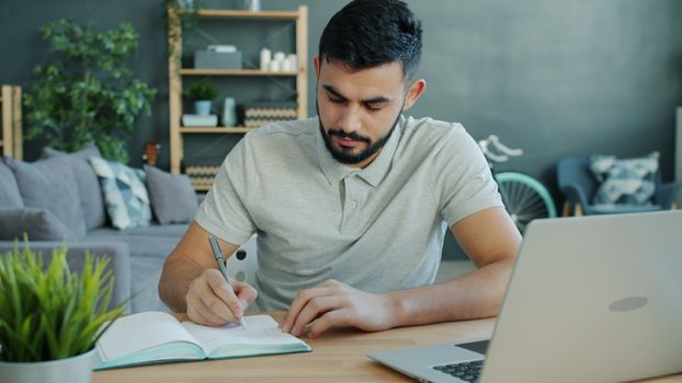 Adult man writing in notebook while working at home with laptop.